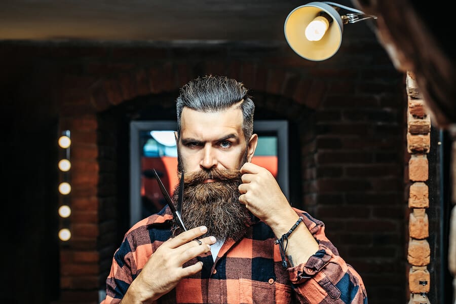Handsome bearded man hipster with stylish haircut and beard holding scissors in red checkered shirt near mirror with serious face in beauty salon or barbershop as barber near brick wall